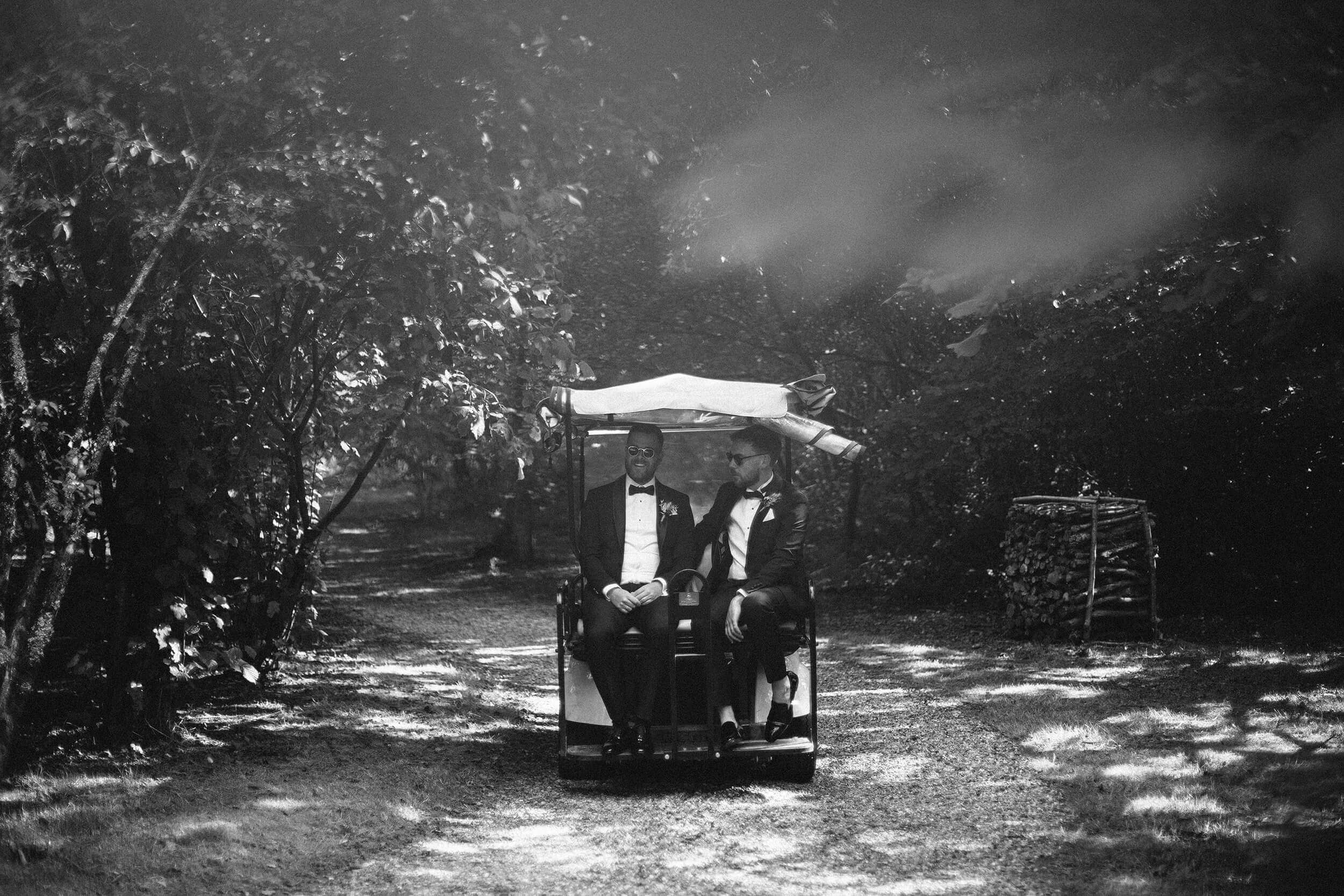 Black and white shot of two grooms laughing together in a golf buggy on a wooded gravel path