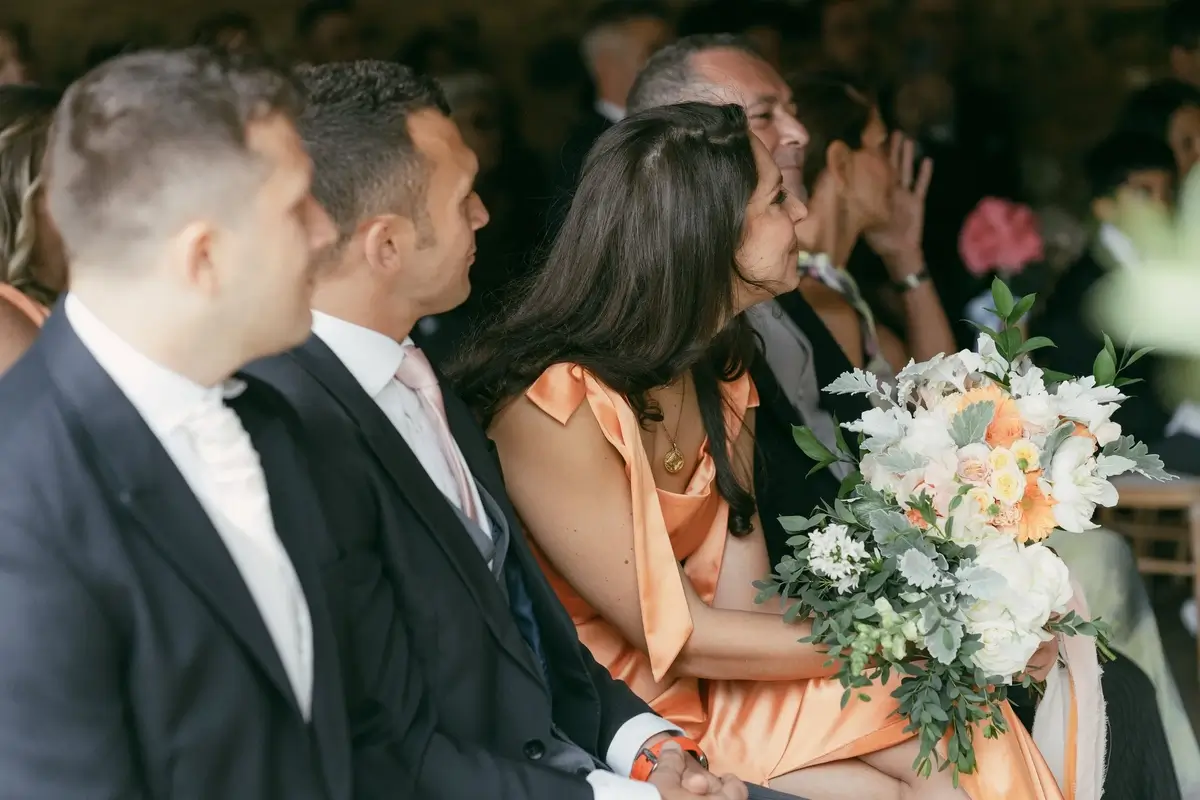 A guest in a coral dress holding the bridal bouquet mid-ceremony as the row around her looks on, smiling