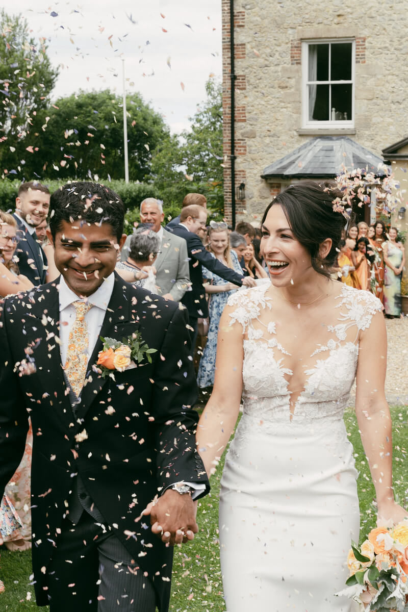 Ashlie and Srini caught mid-laugh in a shower of confetti on the lawn at East Afton Farmhouse