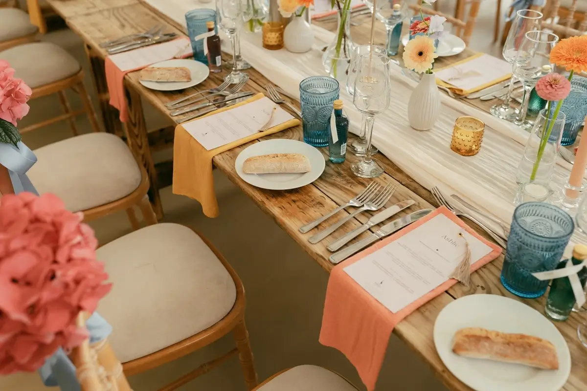 Long feasting tables set with peach and mustard linen, blue glassware and gerberas in bud vases