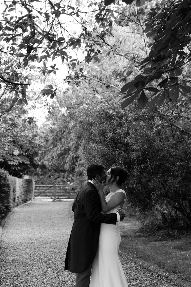 A black-and-white shot of Srini and Ashlie kissing on a tree-lined gravel path, Isle of Wight countryside beyond