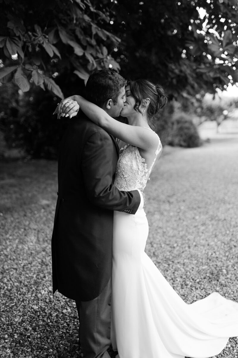Ashlie's hand at Srini's collar as they kiss under chestnut branches, her dress trailing on the gravel