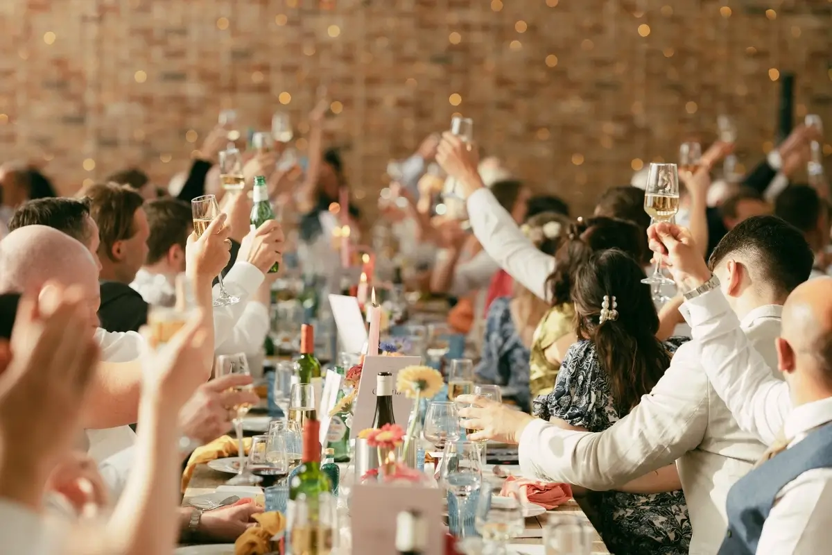 Guests on their feet raising glasses to toast the couple, fairy lights glowing on the brick wall behind