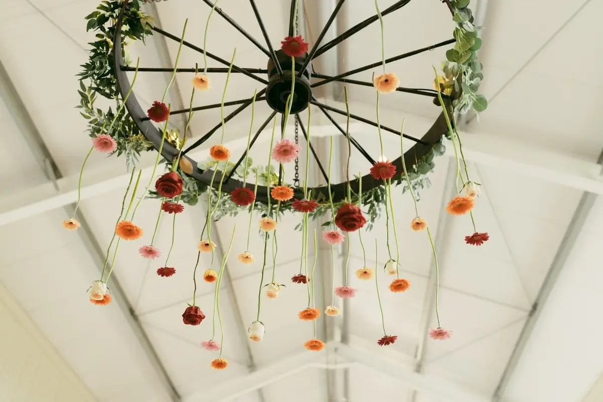 A wagon-wheel chandelier strung with dozens of hanging gerberas and roses in coral, apricot and blush over a barn