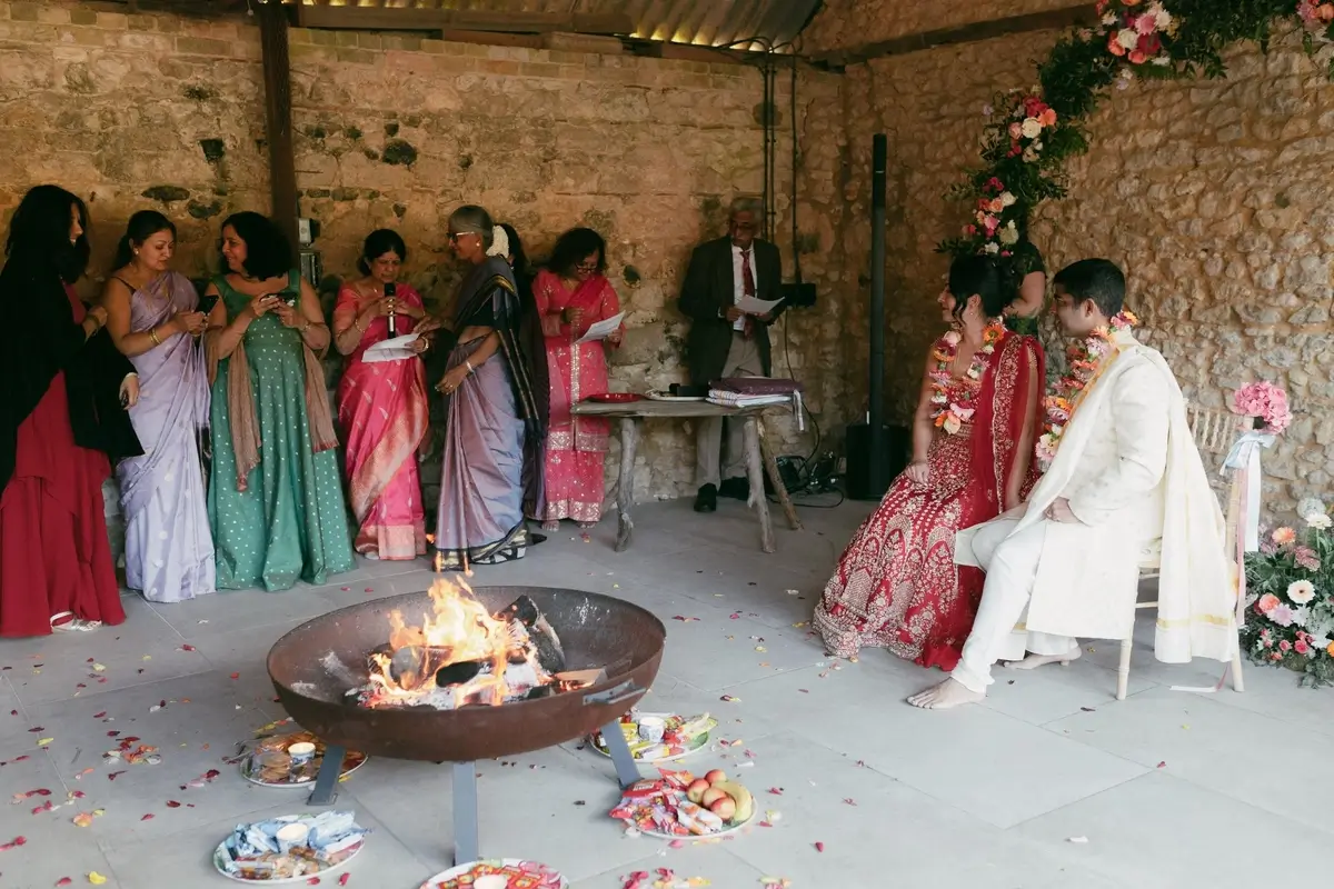 Srini and Ashlie seated by the sacred fire in her red lehenga and his ivory sherwani, family gathered round