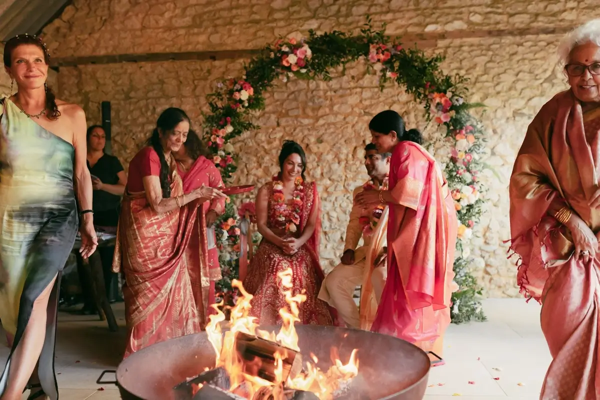 A relative tending the fire during the Hindu ceremony as Ashlie and Srini sit garlanded in coral blooms
