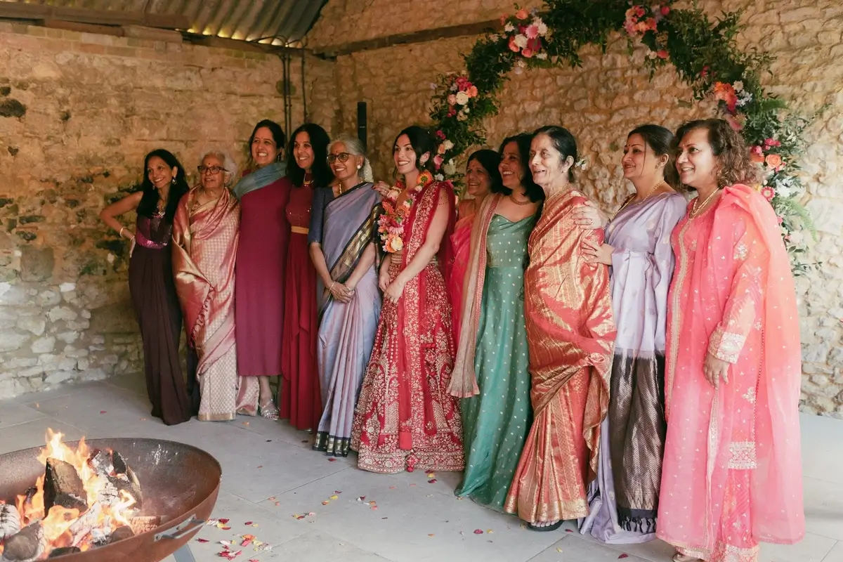 Ashlie in her red-and-gold lehenga surrounded by female relatives in jewel-toned saris by the fire pit