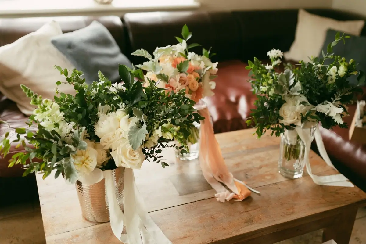 Three bridal party bouquets of ivory roses and greenery lined up on a coffee table, ribbons trailing to the floor