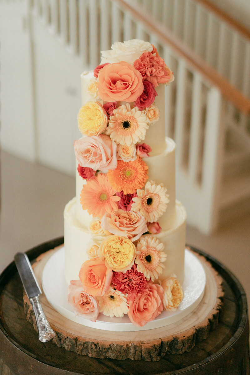 A three-tier buttercream cake heaped with coral roses, gerberas and carnations on a wood-slice stand
