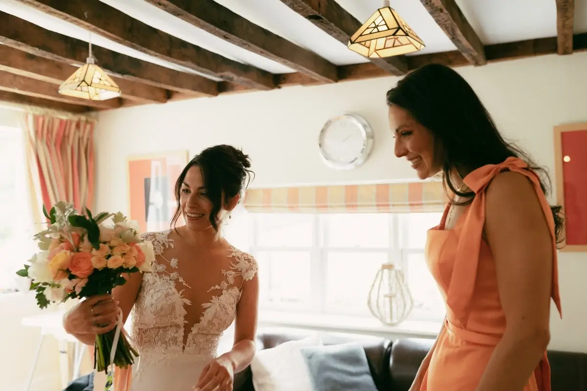 Ashlie laughing with a bridesmaid in a coral dress under the farmhouse's beamed ceiling, bouquet in hand