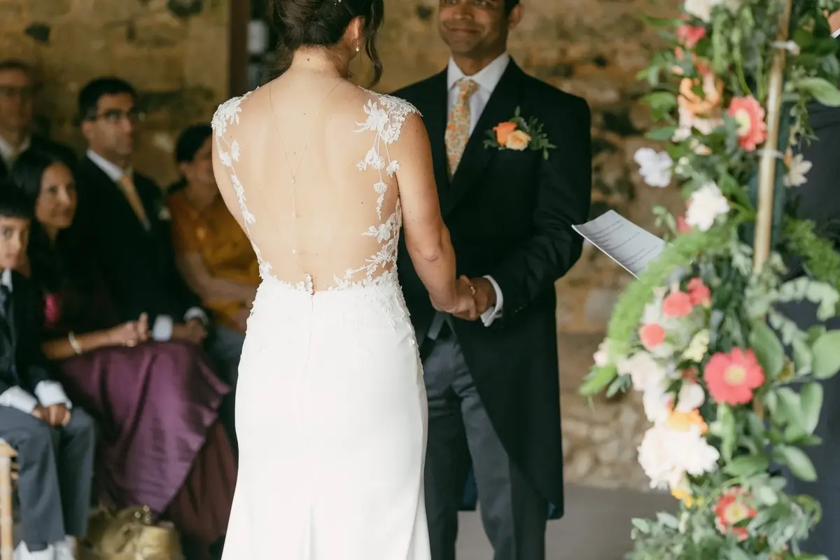 Ashlie and Srini hand in hand at the altar, her open lace back to camera, coral gerberas banked beside them