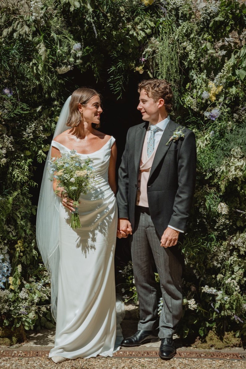 Cesca & Seb hand in hand beneath a tumbling arch of wild garden flowers at the church door