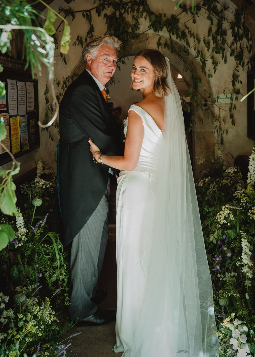 Bride smiles back over her shoulder on her father's arm in the flower-framed church doorway