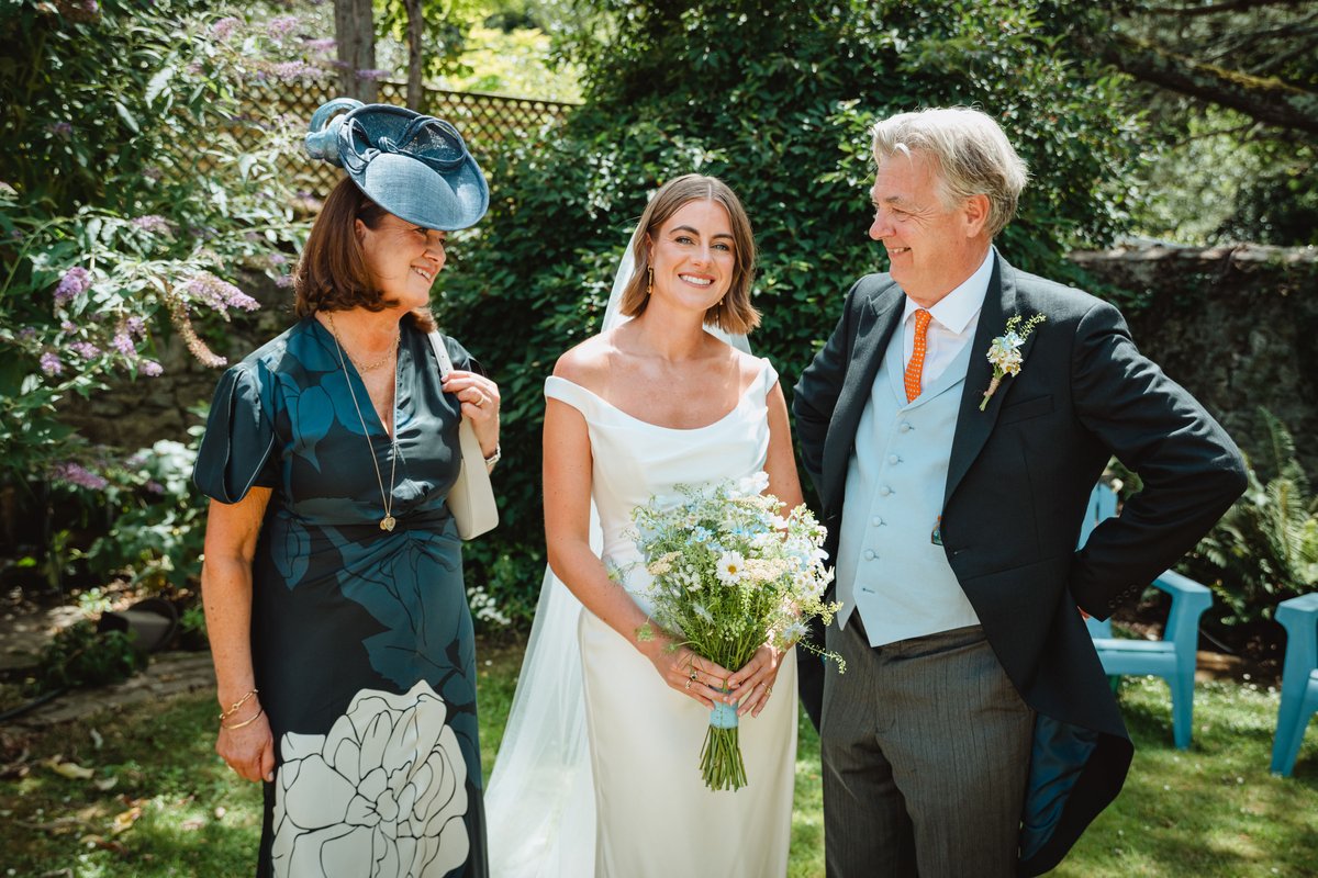 Bride holding her wildflower bouquet and sharing a laugh with her parents in a sunny garden