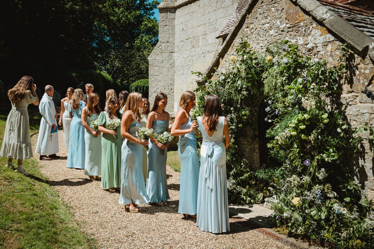 Bridesmaids in blue and sage slip dresses queue at the church porch beneath wild garden flowers