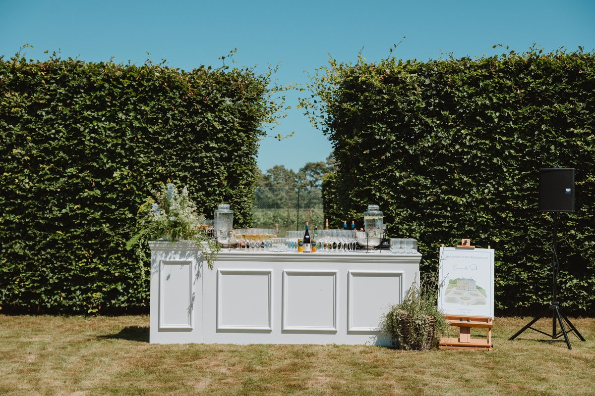 Panelled white bar with drinks dispensers and a welcome sign set against tall beech hedges