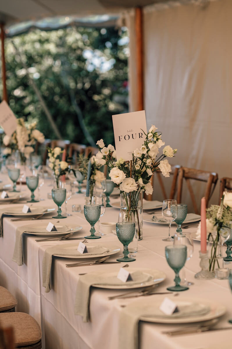 Long reception table under the sailcloth tent with green pressed-glass goblets, coral candles and white florals