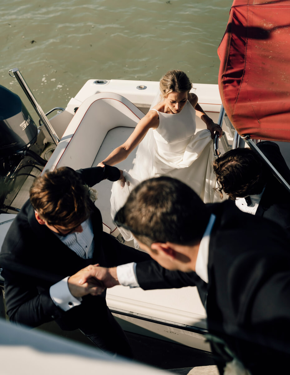 Bride in white dress stepping onto boat as two groomsmen offer steadying hands