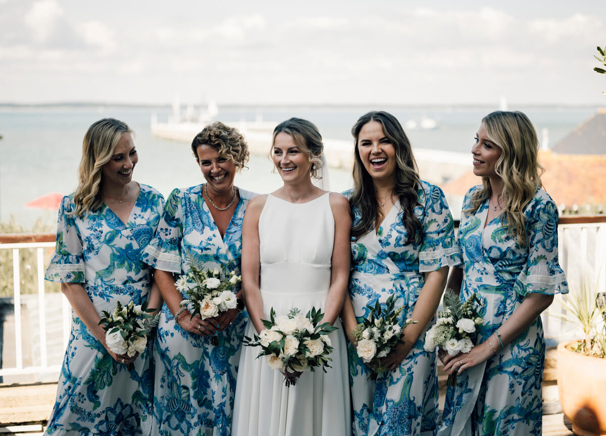 Bride and four bridesmaids in blue floral dresses laughing together by the Solent