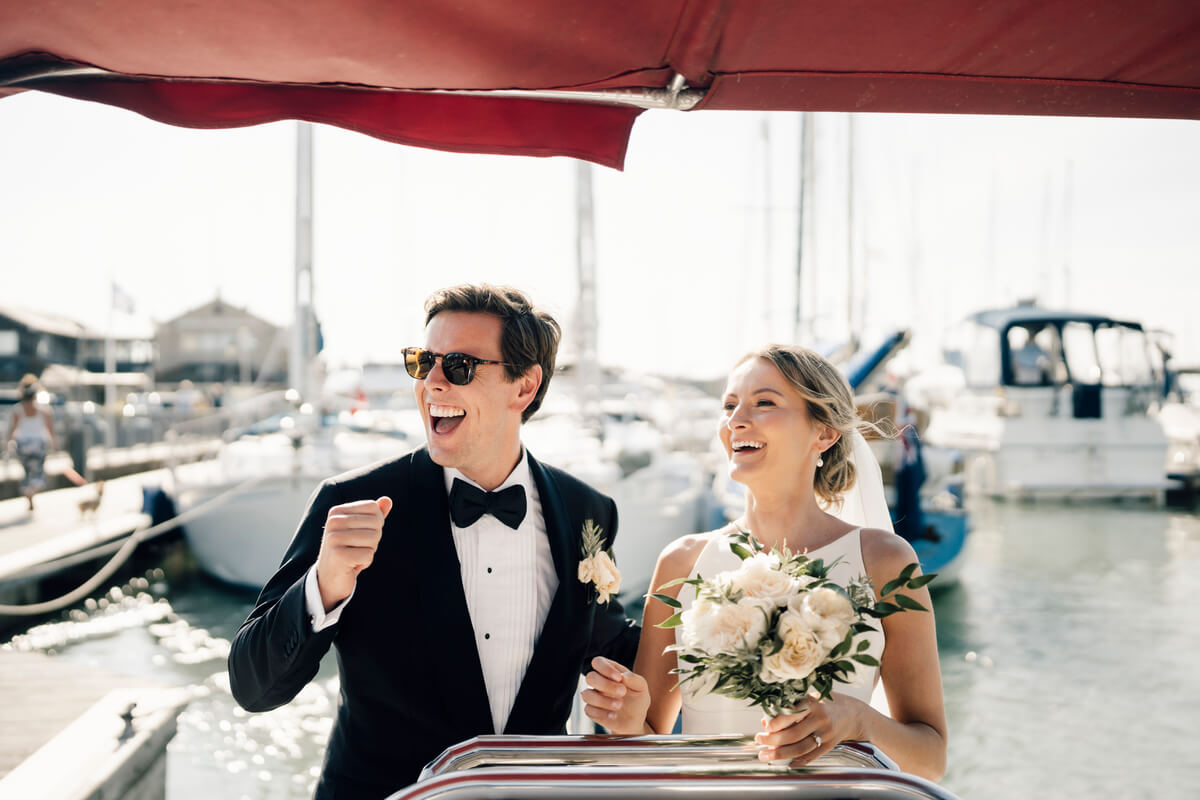 Bride and groom laughing aboard a boat in Yarmouth harbour, white rose bouquet in hand