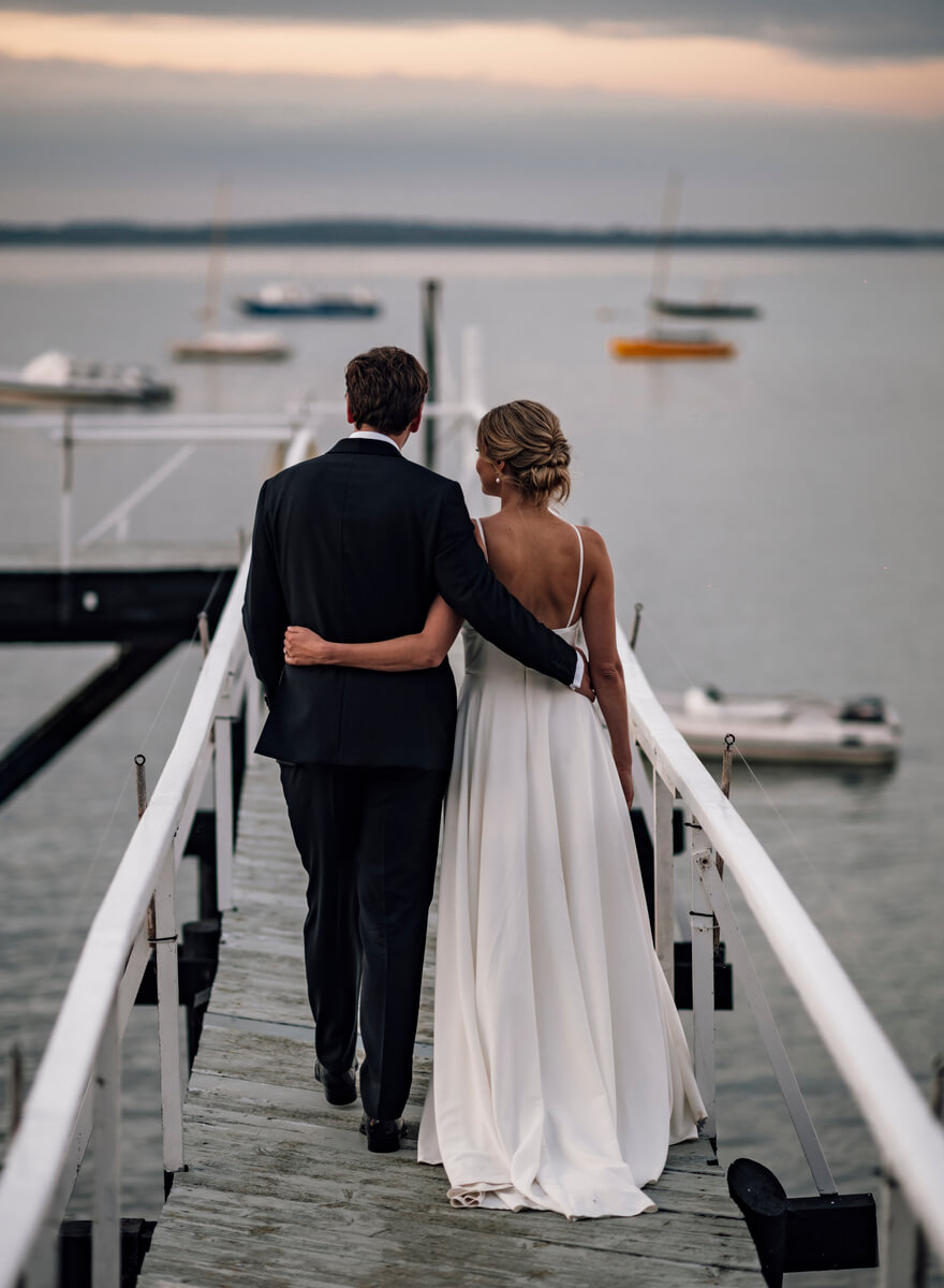 Bride and groom walking arm in arm along a white wooden jetty at dusk over the Solent