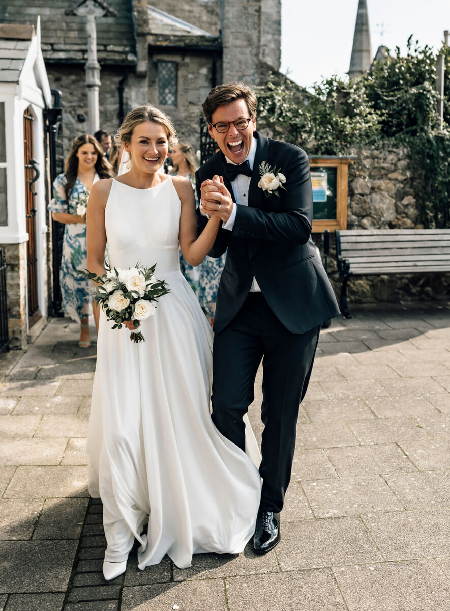 Bride and groom laughing as they walk hand in hand from a stone church on a sunny day