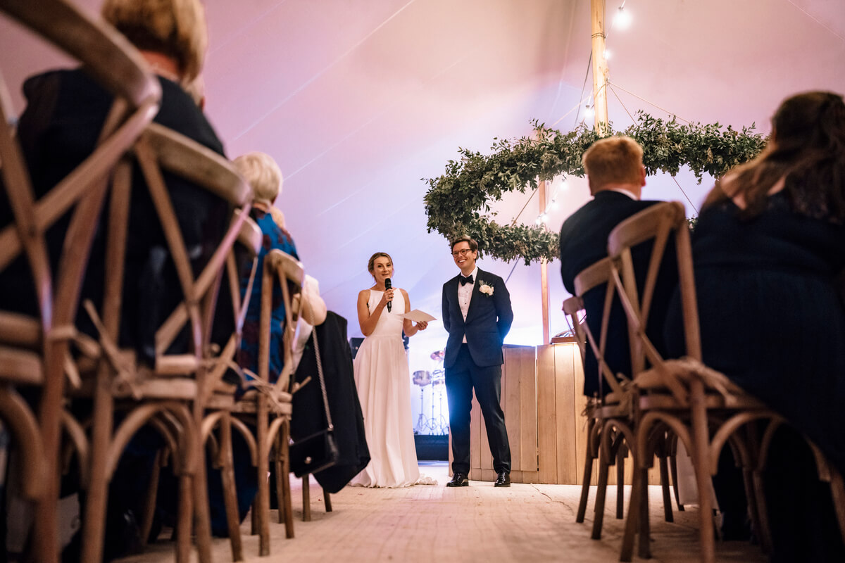 Bride giving a speech beside the groom under a green foliage arch inside the sailcloth tent
