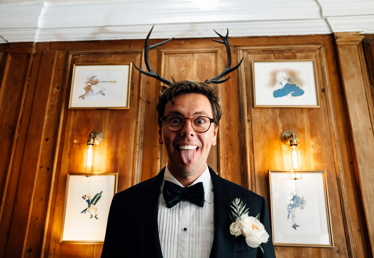 Groom in tuxedo pulls a face with mounted antlers framing his head in a panelled room
