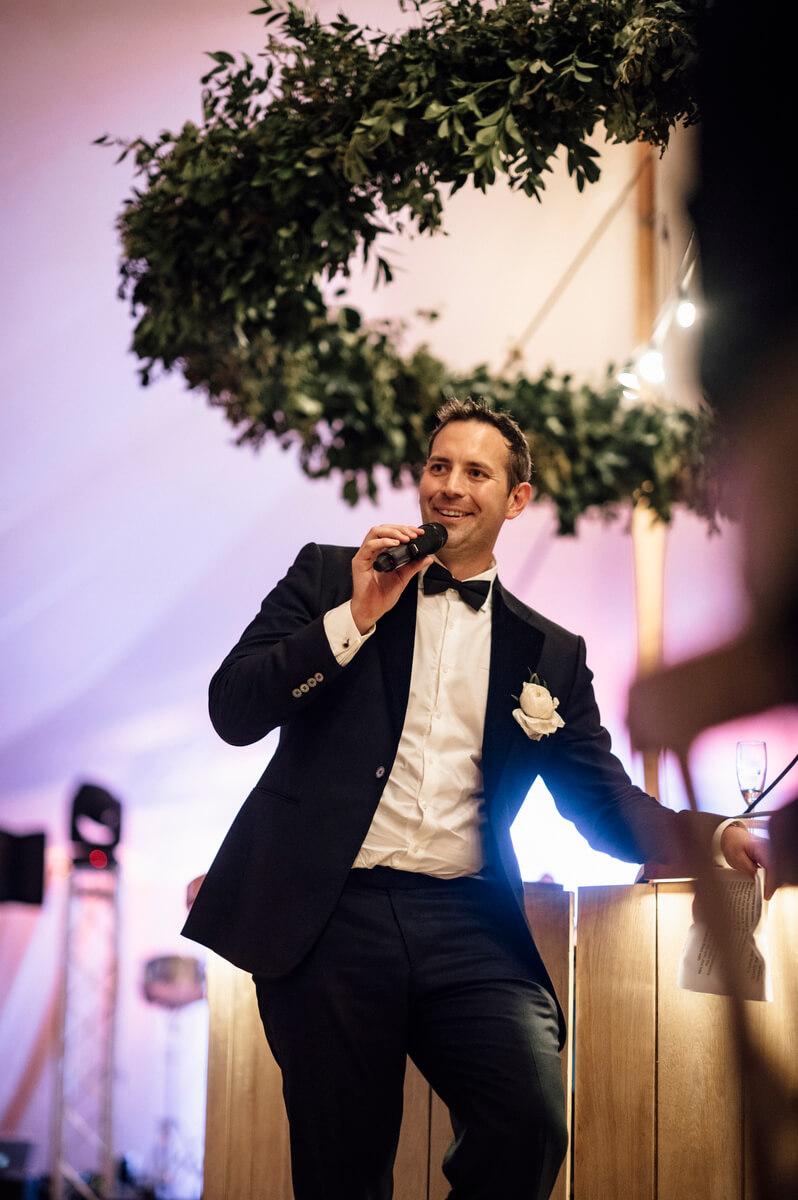 Groom smiling mid-speech with microphone in hand under a foliage hoop in the tent