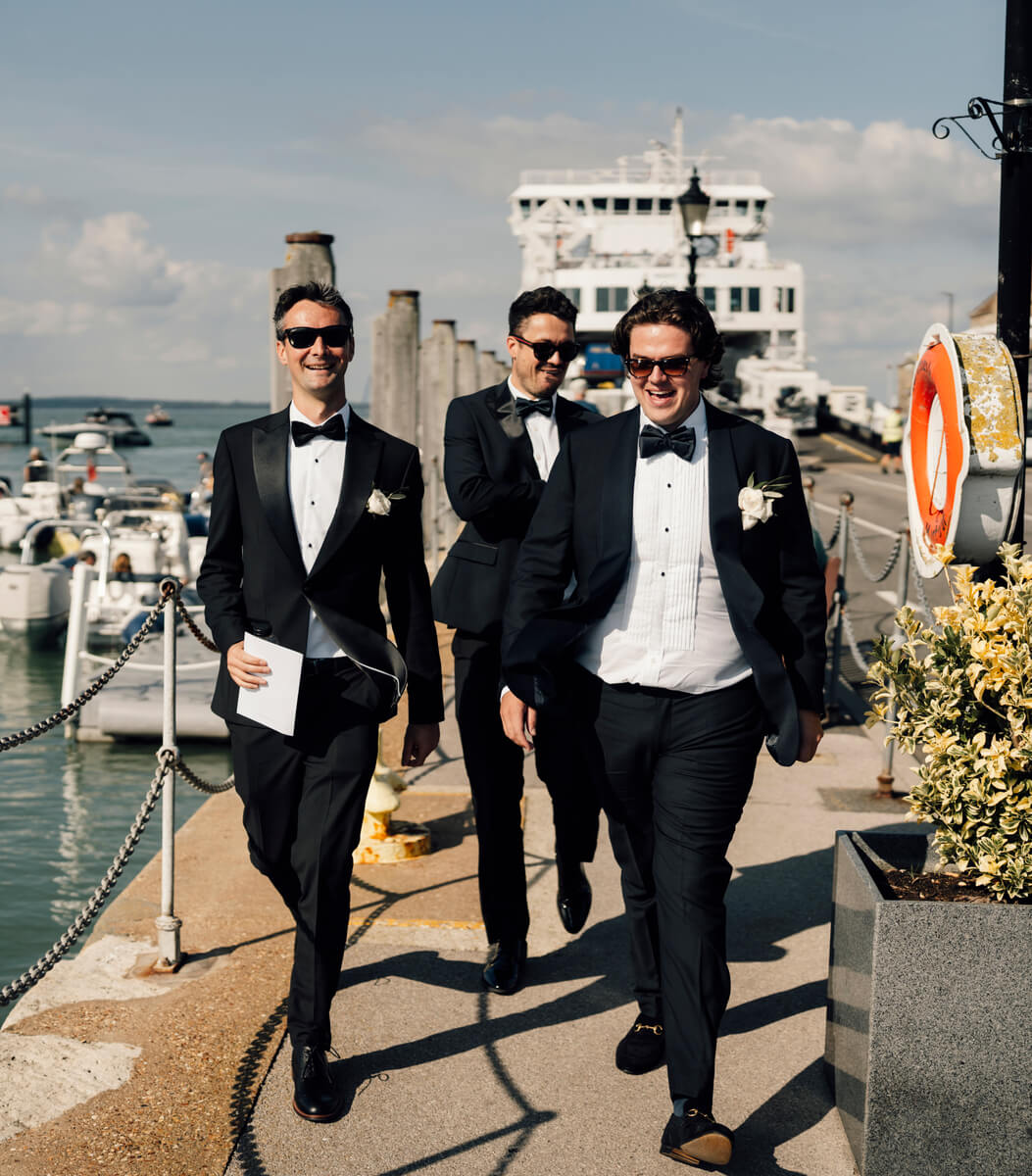 Three groomsmen in tuxedos walk along Yarmouth harbour with the Wightlink ferry behind