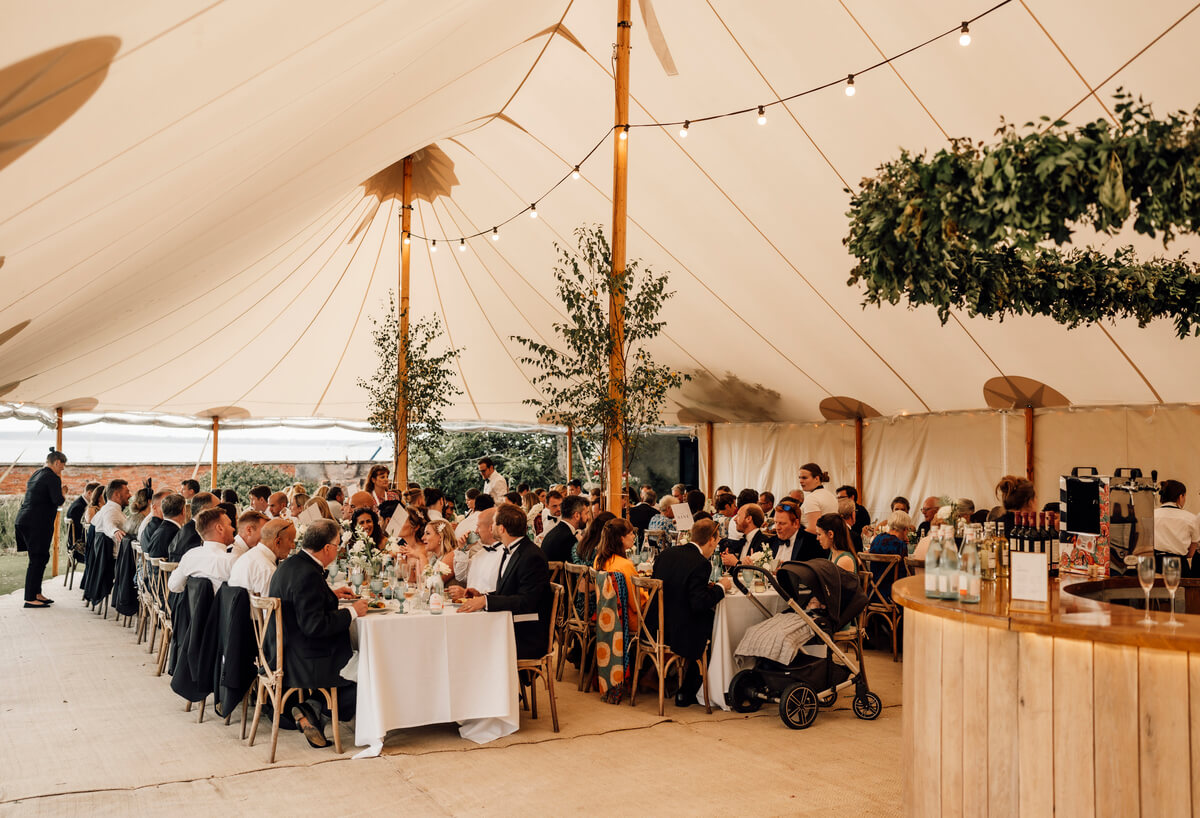 Guests seated for the wedding breakfast at long tables under a sailcloth tent with hanging foliage