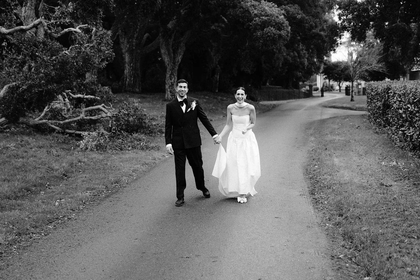 Black and white shot of Liam leading Fiona by the hand along a tree-lined path at Barton Manor, both laughing