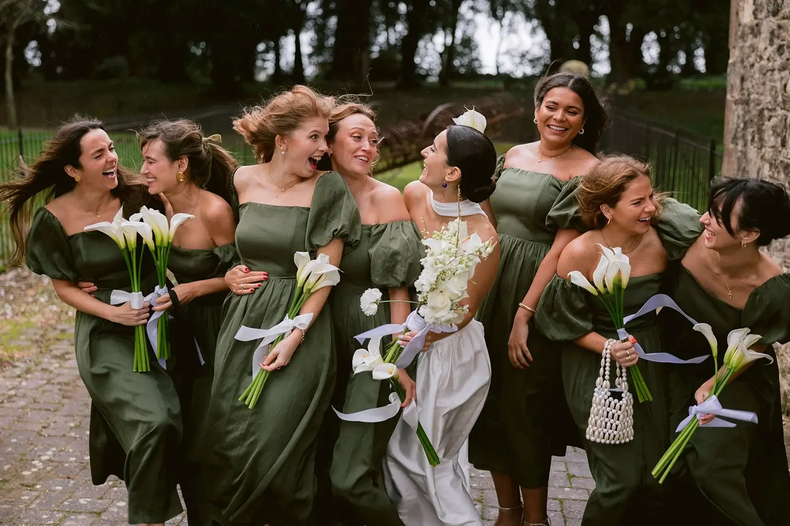 Fiona and her bridesmaids in olive green dresses laughing together, clutching white calla lily bouquets