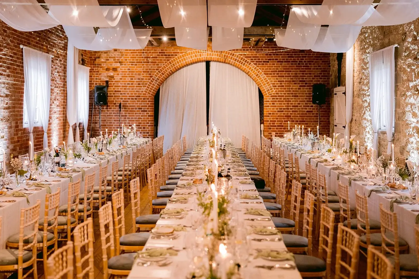 Long feasting tables dressed in white linen inside a brick barn, candlelight and fairy-lit drapes overhead