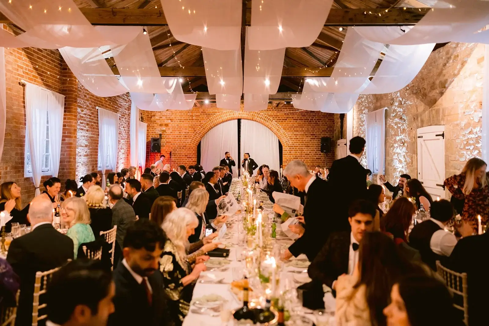 Wide view of the reception in full swing, long guest tables filling the brick barn under white drapes