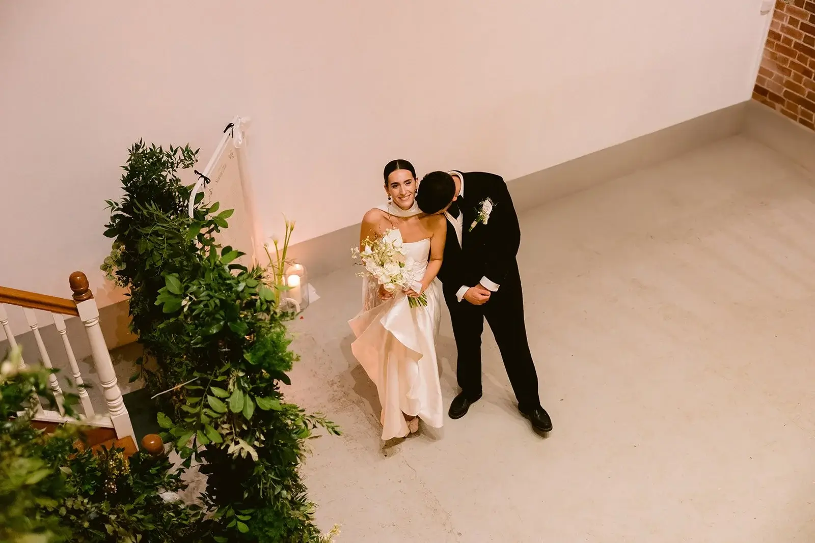 Overhead shot of Liam kissing Fiona's cheek beside a greenery-wrapped staircase