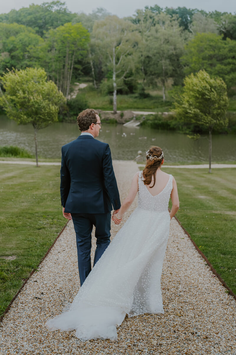 Bride and groom holding hands walking down a gravel path toward the pond at Barton Manor