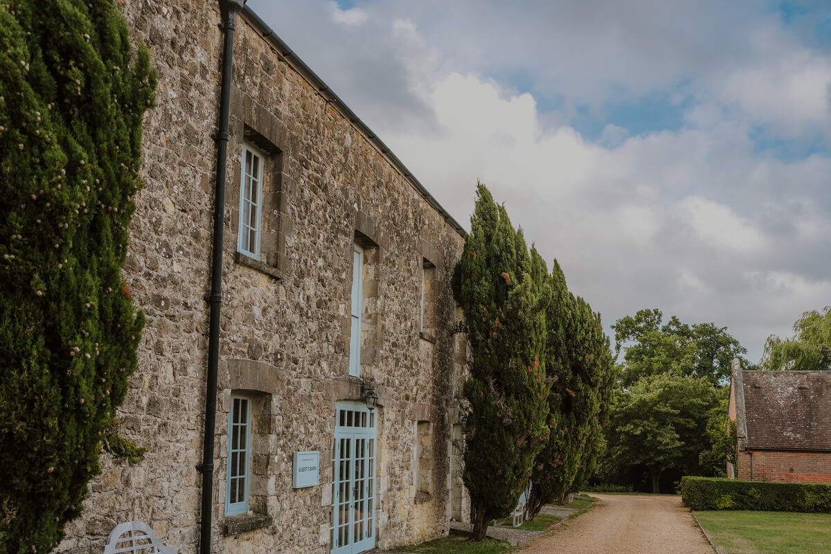 Sunlit stone barn exterior lined with tall cypress trees at Barton Manor