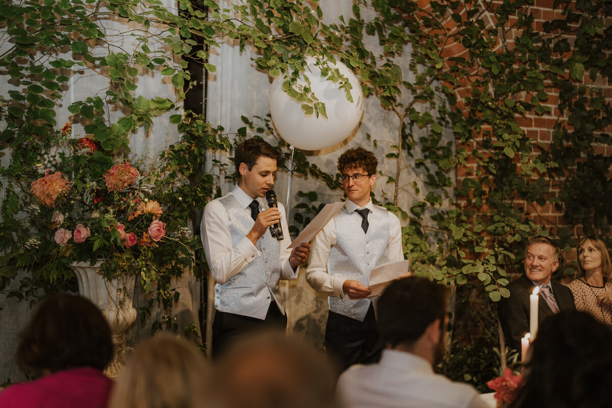 Two best men giving a speech in front of a leafy white moongate arch