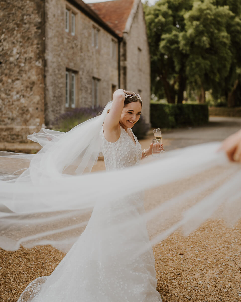 Laughing bride twirls her sparkling veil while holding champagne outside a stone manor house
