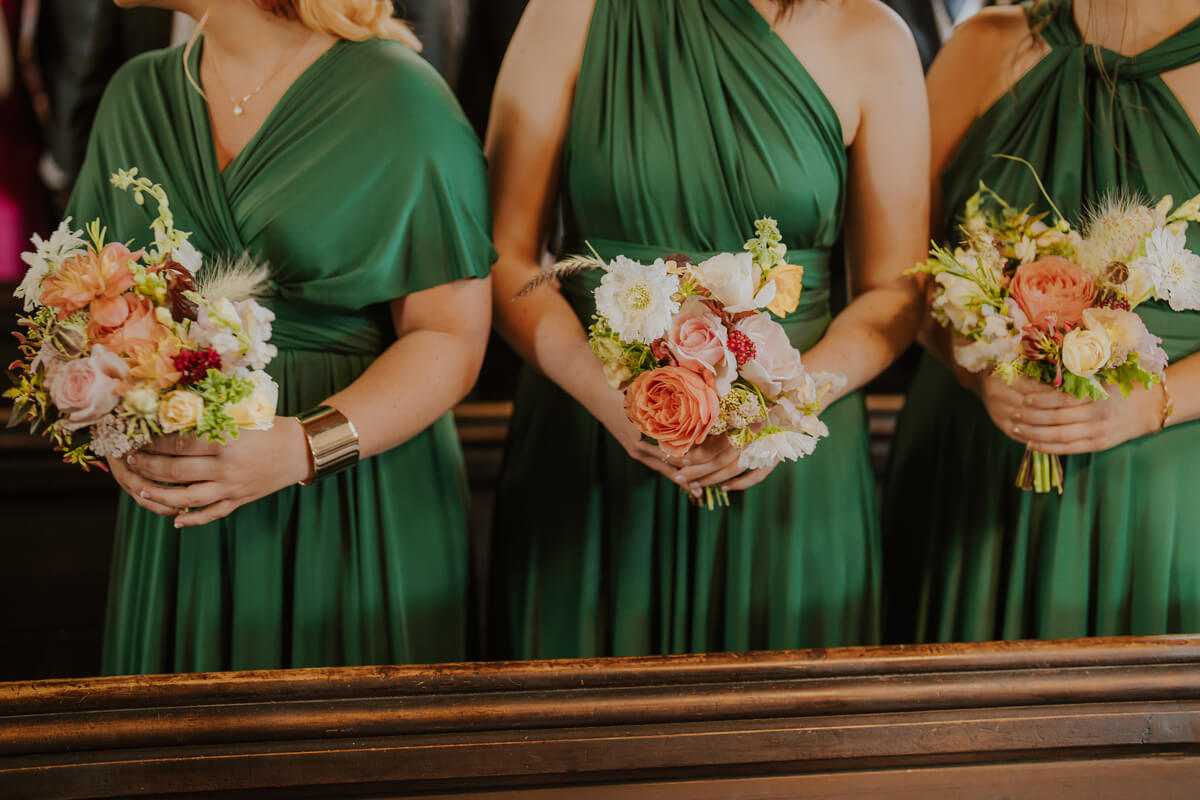 Three bridesmaids in emerald green dresses holding peach and coral pastel garden bouquets