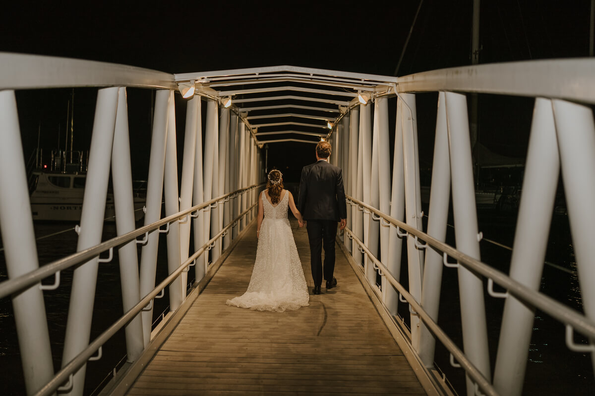 Bride and groom walking hand in hand down a lit marina gangway at night in Cowes