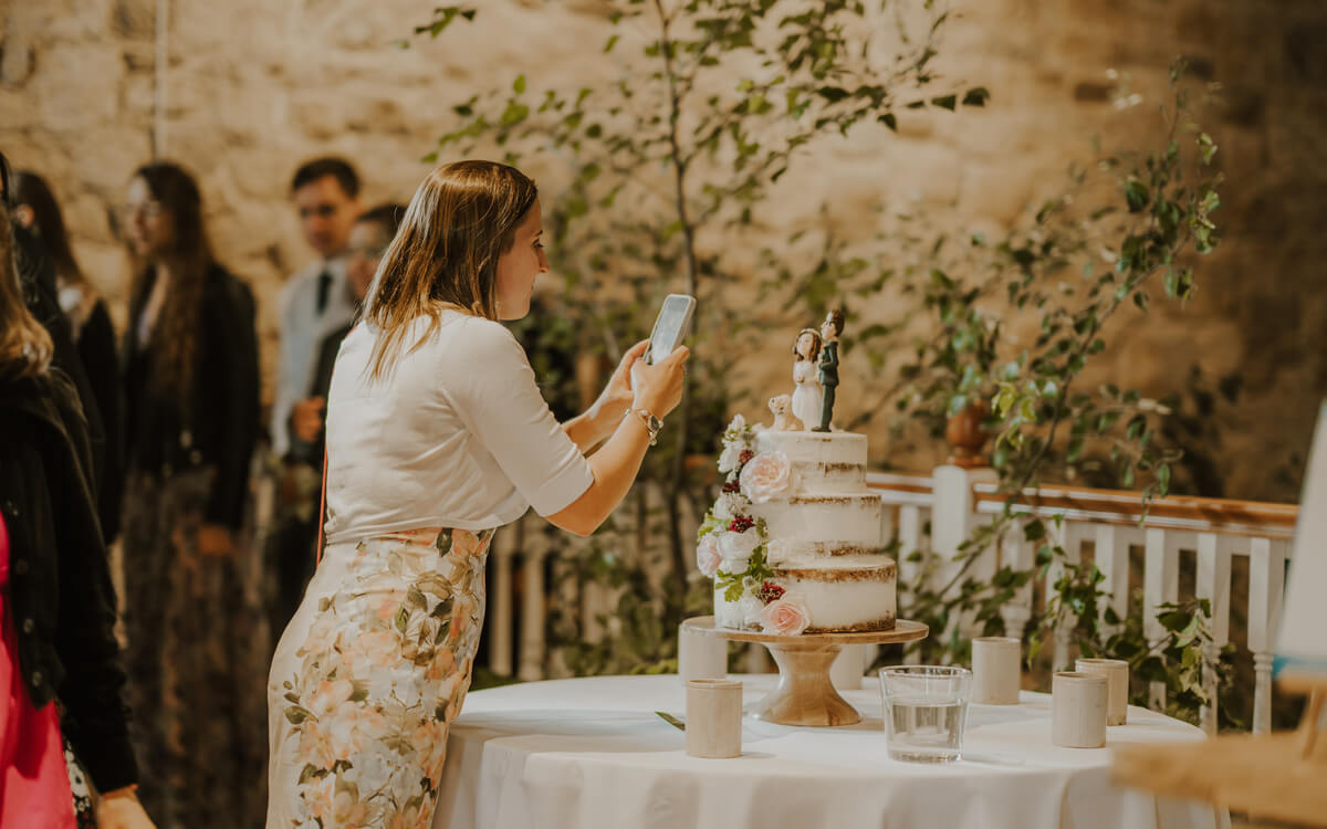 Wedding guest photographing a semi-naked tiered cake with hand-painted bride and groom topper