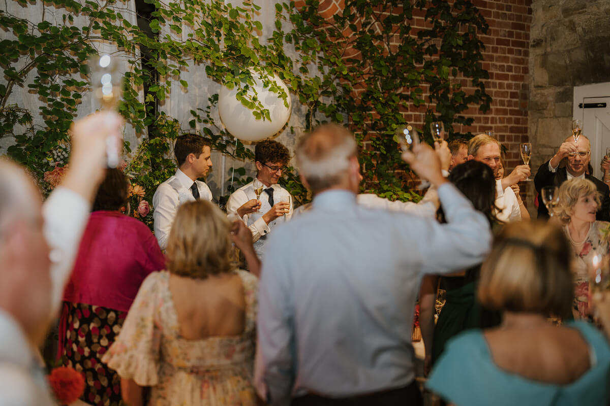 Reception guests raising champagne glasses to toast two groomsmen during their speech