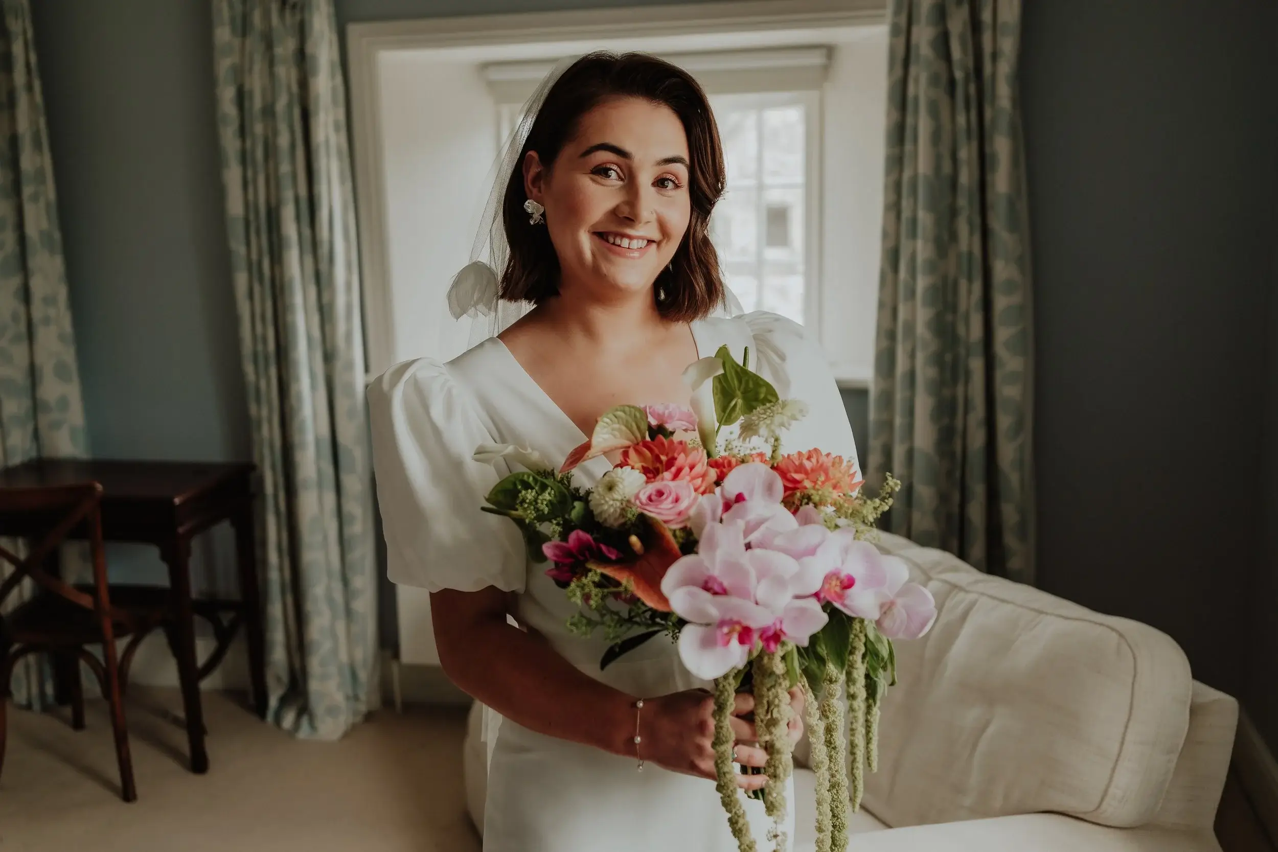 Georgina smiling by a sash window holding her bouquet of orchids, dahlias and anthurium before the ceremony