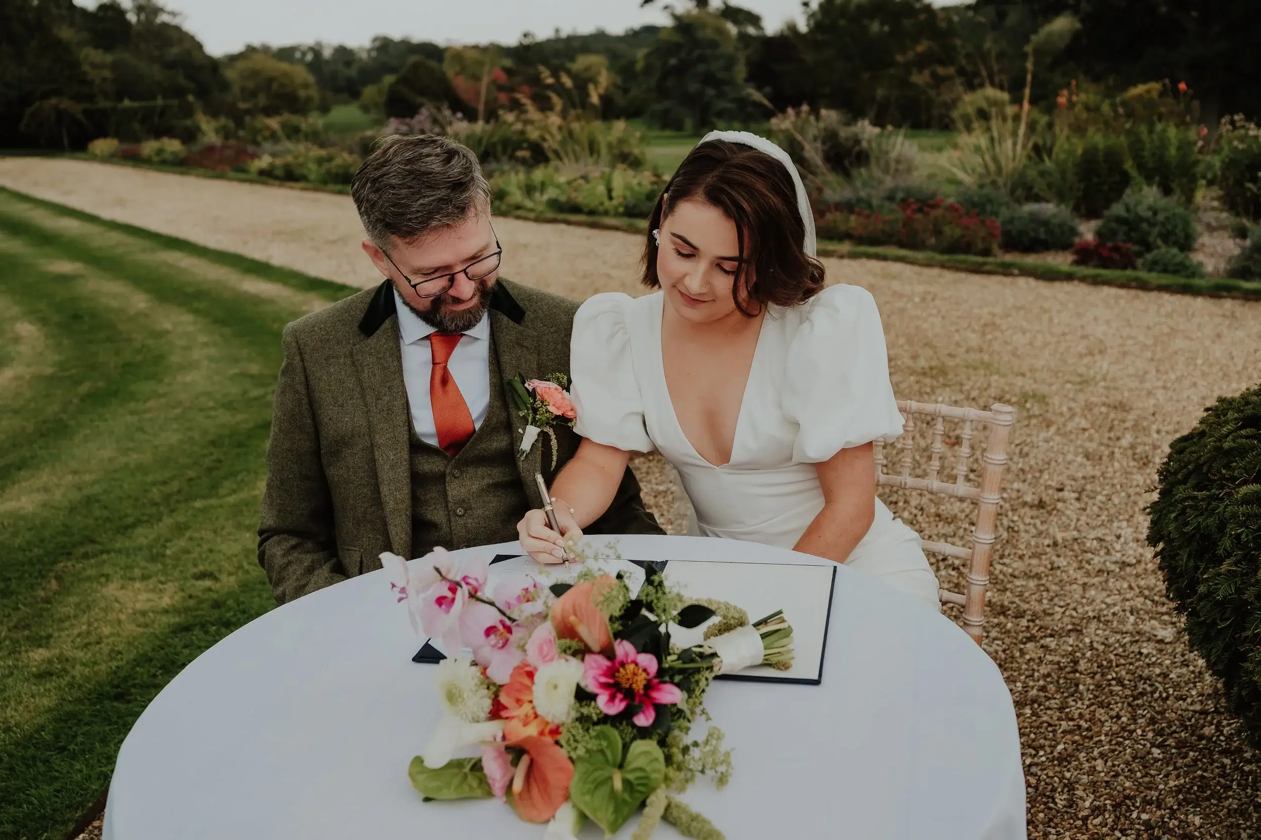 Georgina and Liam signing the register together at a table topped with their bouquet, gardens rolling out behind them