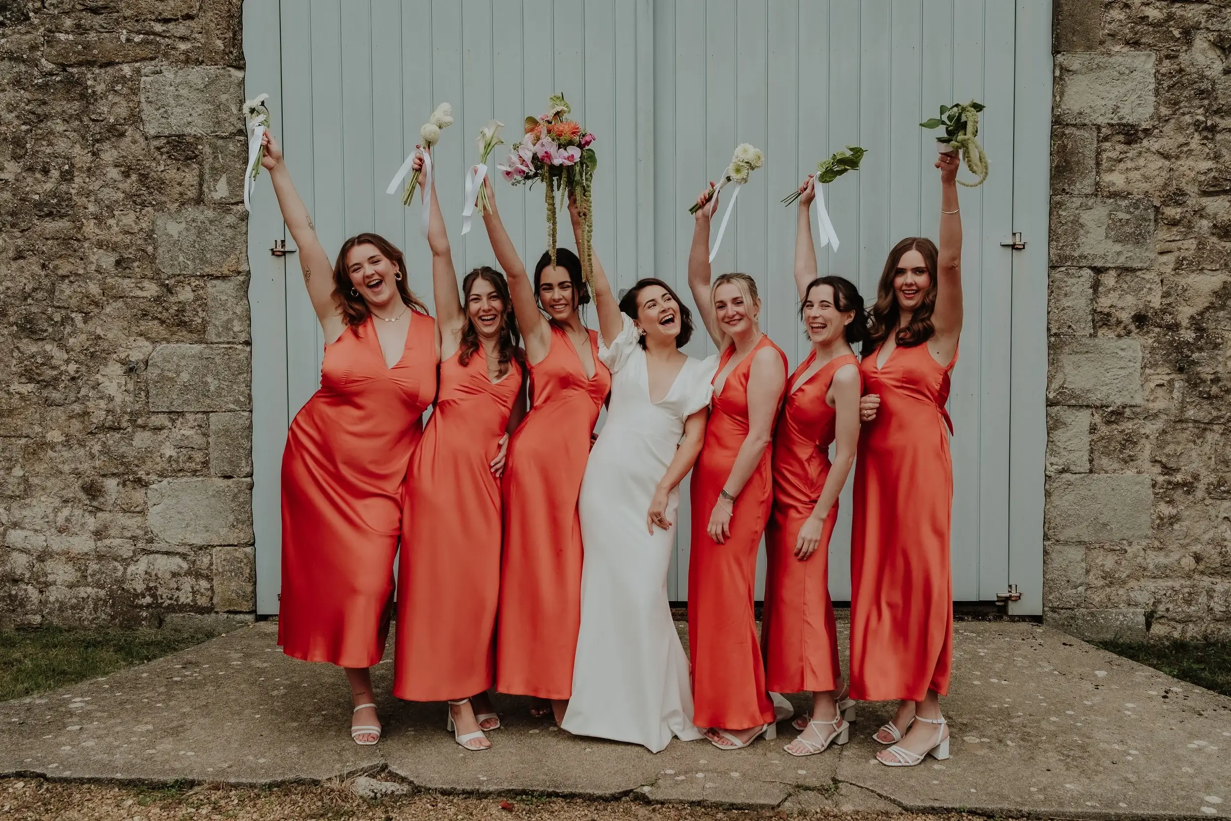 Georgina and her bridesmaids in orange satin dresses cheering with bouquets raised outside a pale barn door