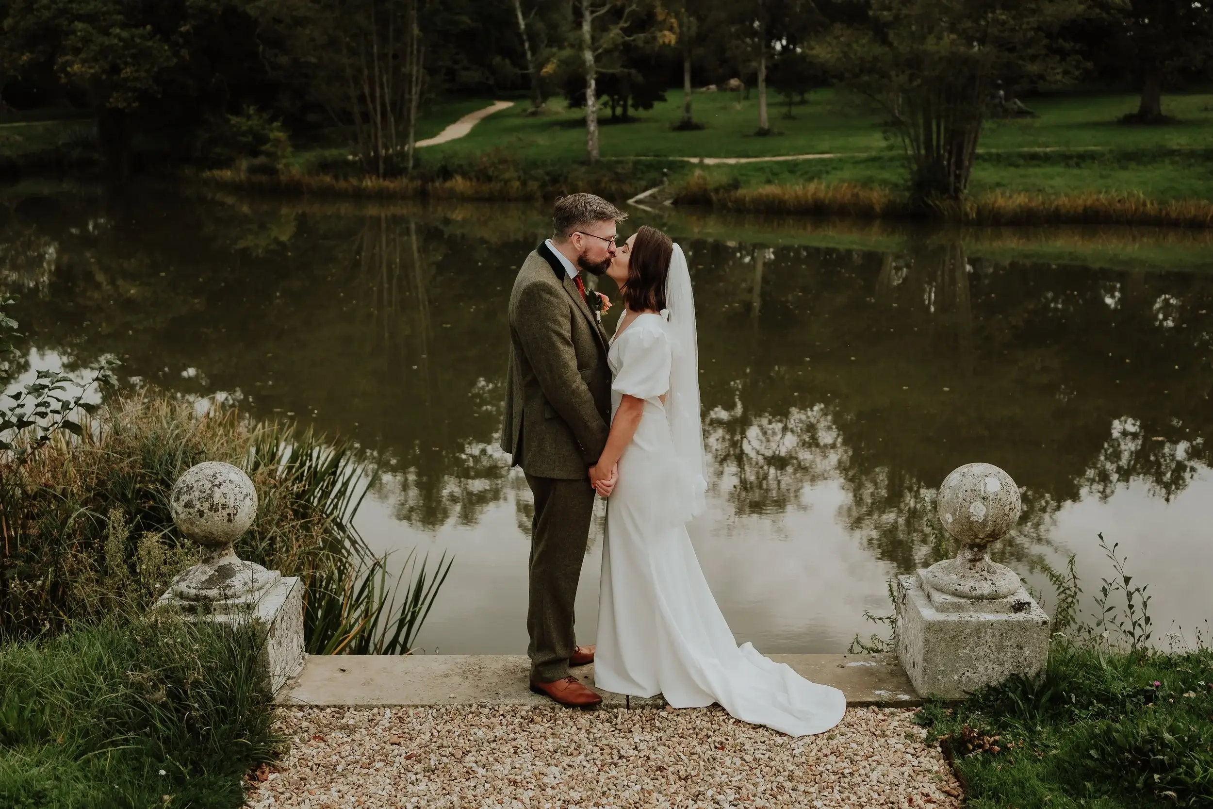 Georgina and Liam sharing a kiss by the lake's stone balustrade, the water and autumn trees reflected behind them