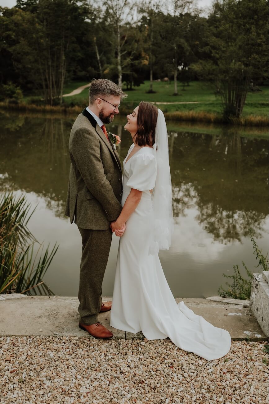 Georgina and Liam smiling at each other on the same lakeside terrace, her veil catching the breeze
