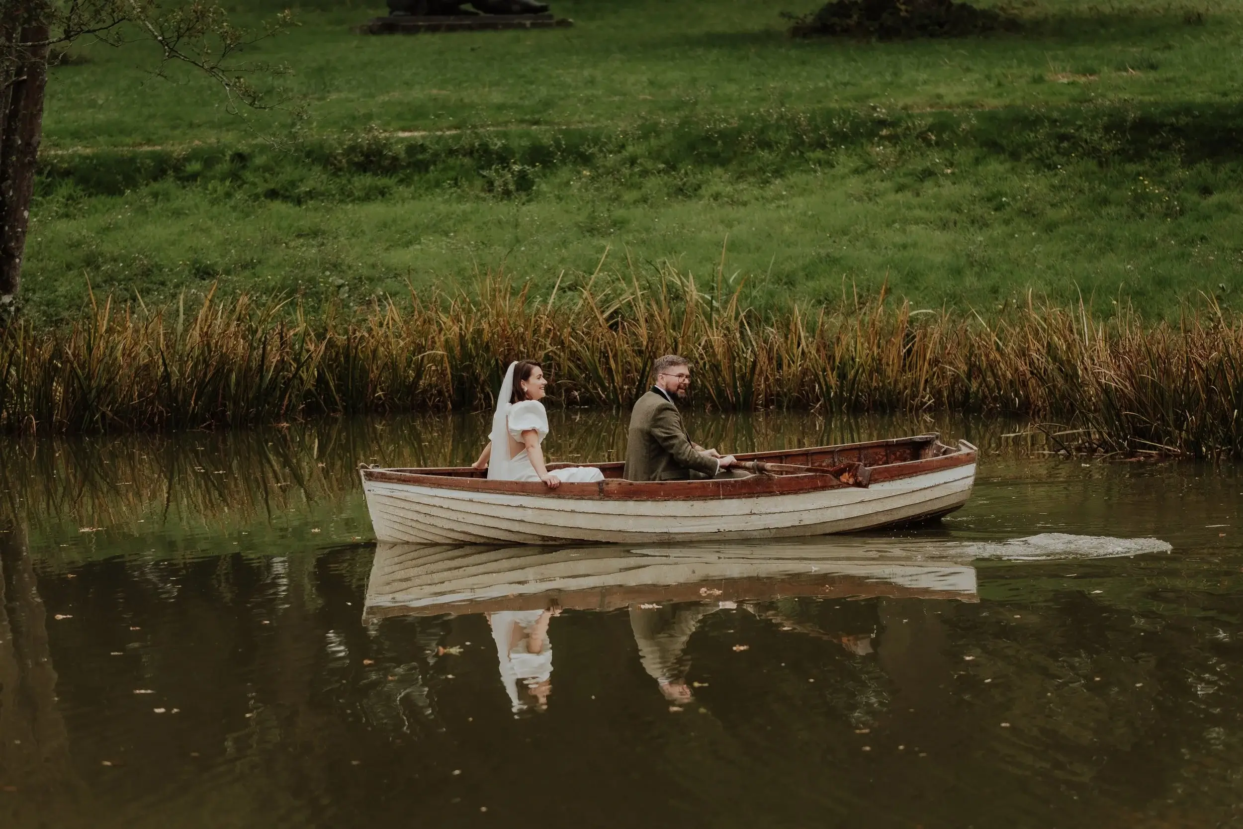 Georgina and Liam rowing across the lake in a wooden boat, reeds and open lawn along the far bank